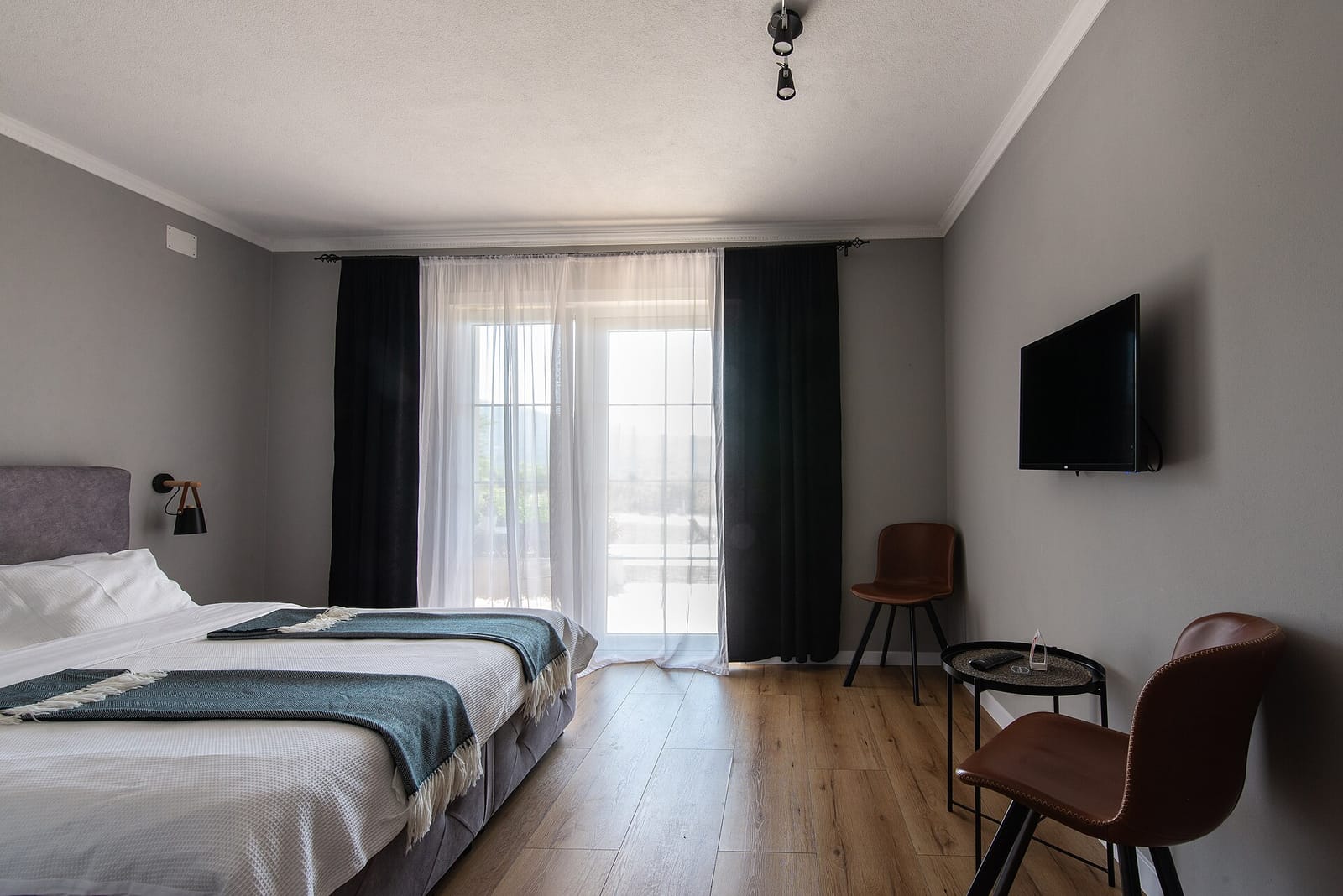 A freshly made double bed with white bedding and a folded throw, beside a wide window with a sheer curtain drawn back.