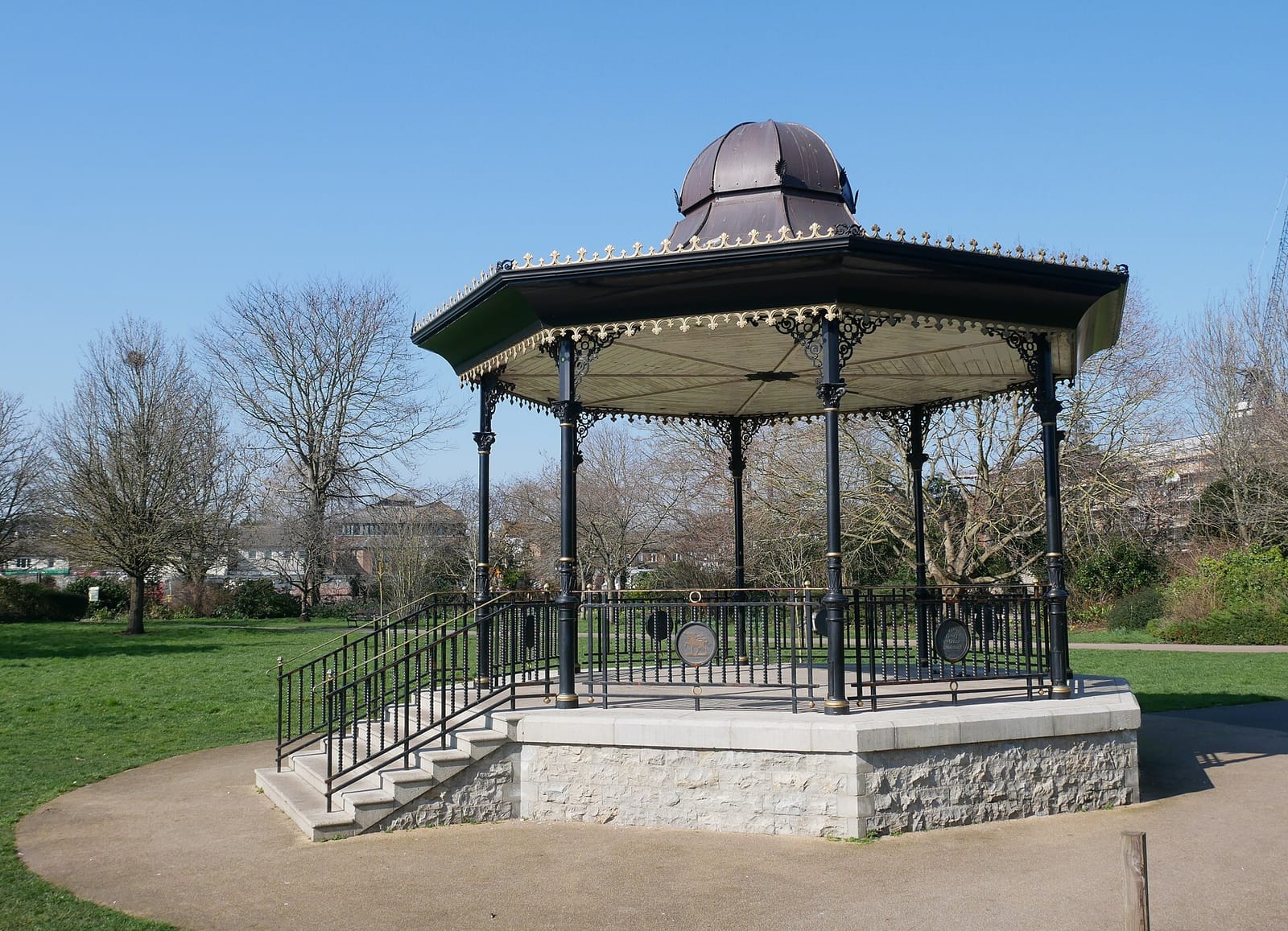The restored Victorian bandstand in Central Park, Dartford, standing on its stone base under a clear blue sky.