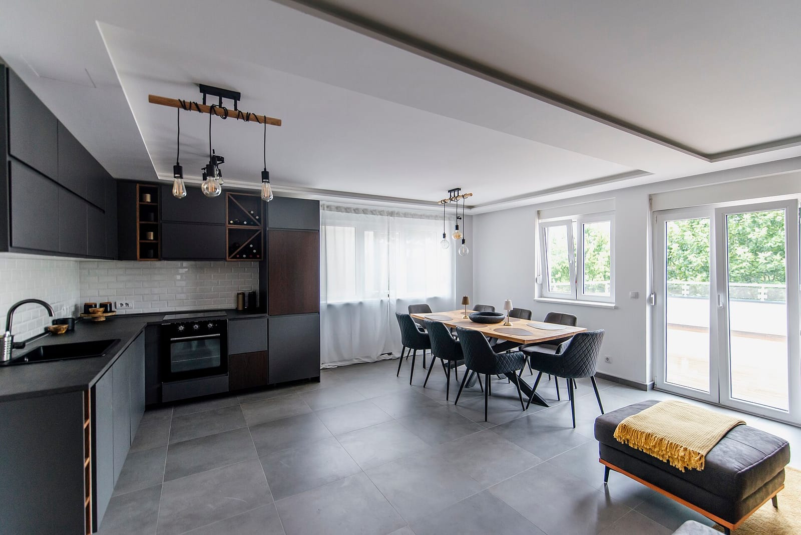 A bright open-plan kitchen and dining space with dark grey units, white metro tiling and a wooden table laid under tall windows.